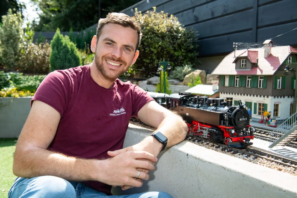 Curtis Stanier smiling beside the HSBA Garden Railway, with a steam locomotive on the track behind him